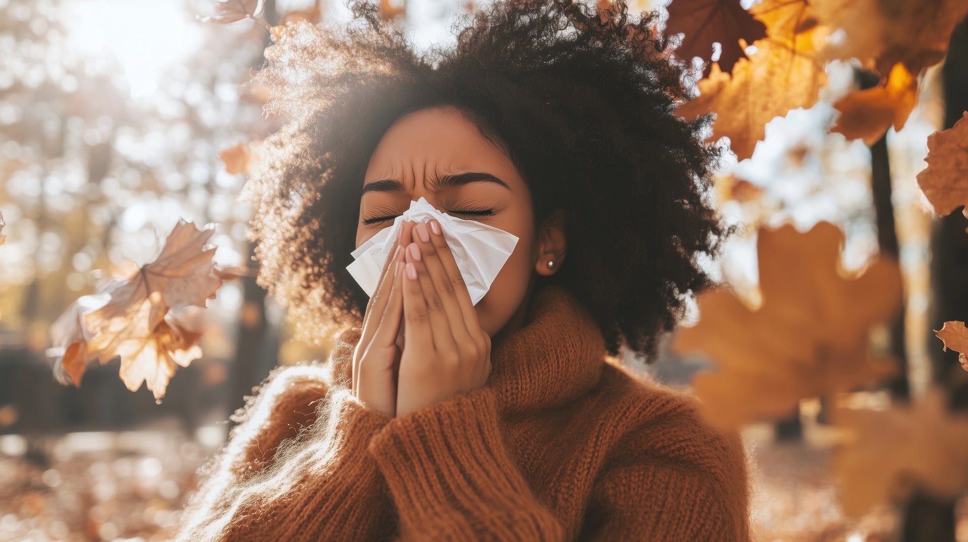 Woman sneezing in autumn leaves, experiencing allergies, wearing cozy sweater