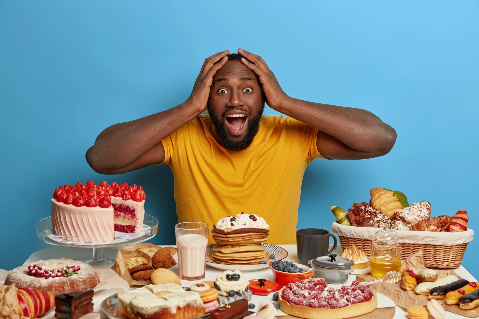 Man with both hands on head, opens mouth looking at a table of sugary desserts