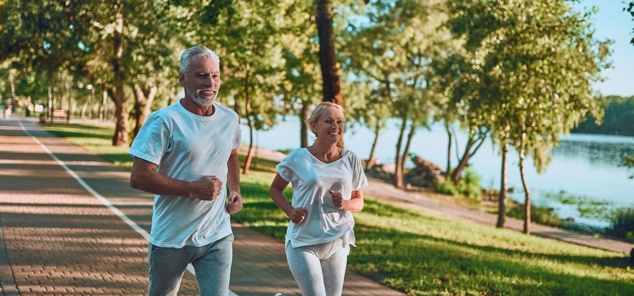 Older man and woman running alongside lake with green trees.