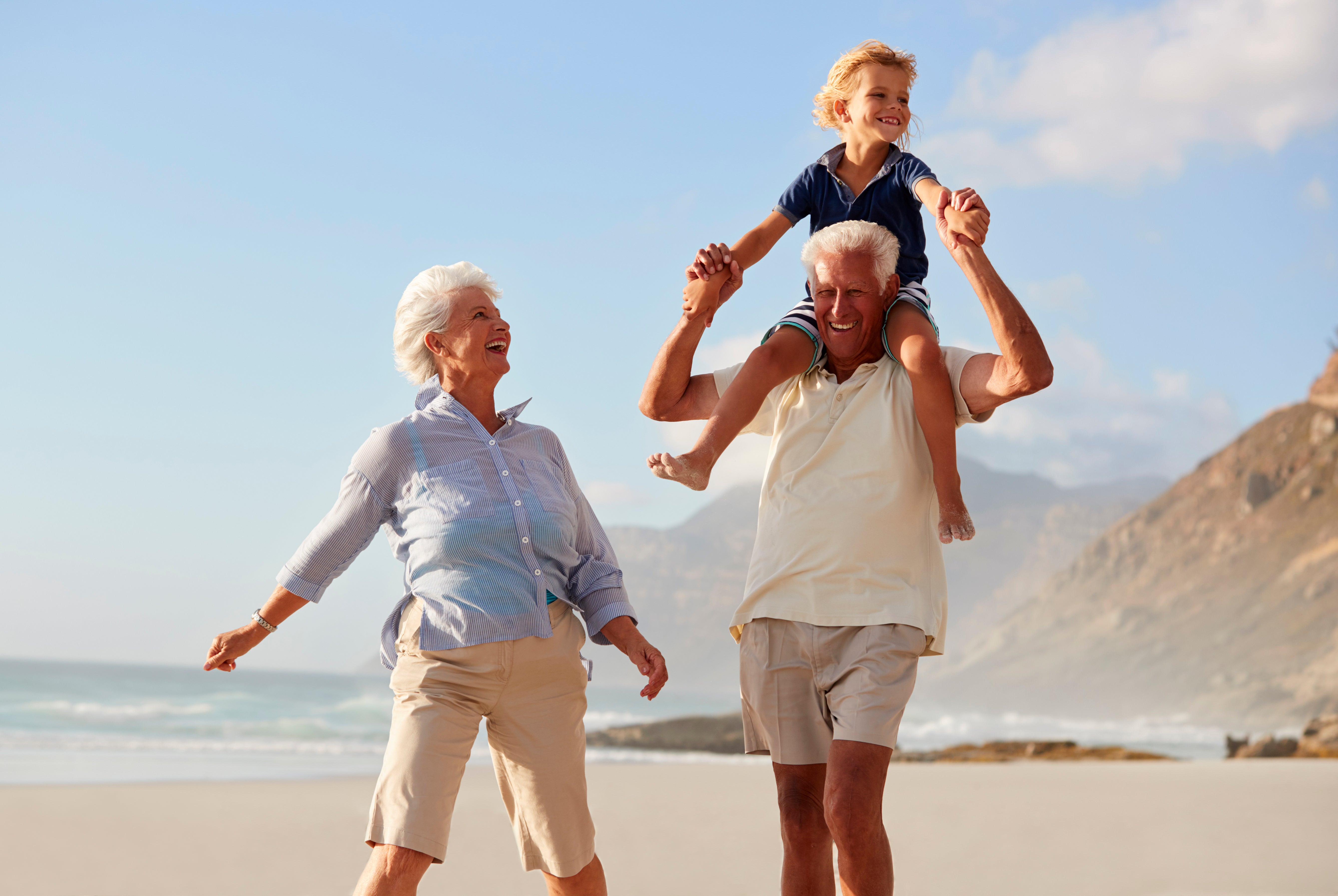 Older Adults Carrying Grandson On Shoulders On Walk Along Beach.
